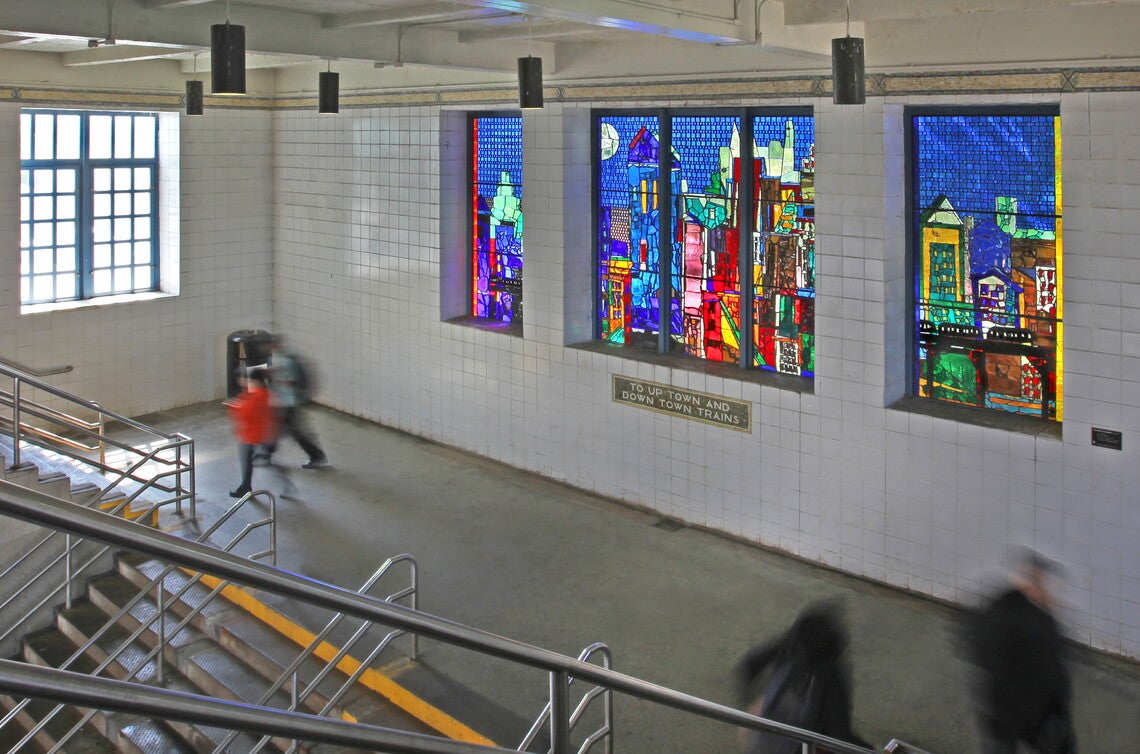 A color photograph of people at right foreground and left background going up a set of stairs; in the center background, a multicolored glass mosaic of a city skyline with an elevated train, and below a tile mosaic reading "TO UP TOWN AND DOWN TOWN TRAINS."