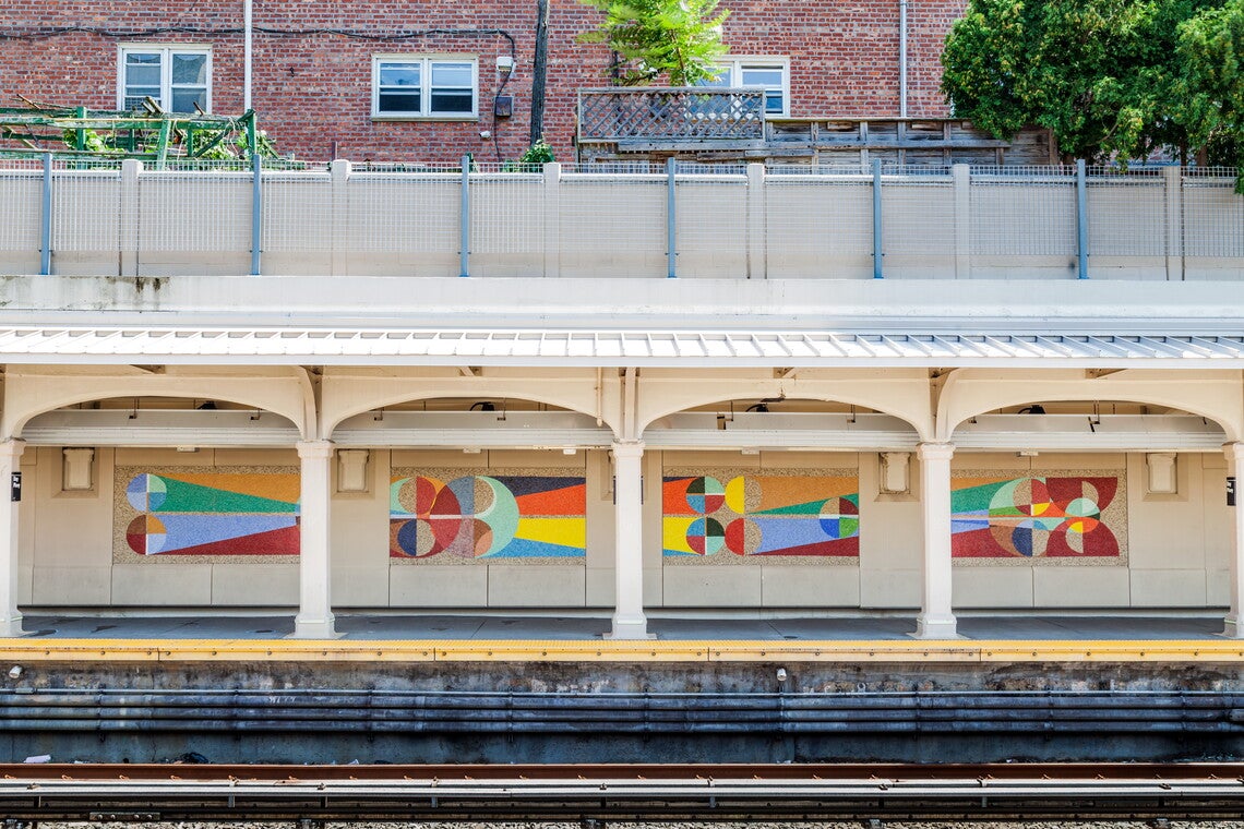 A color photograph of an outdoor subway platform with four multicolored abstract mosaic panels