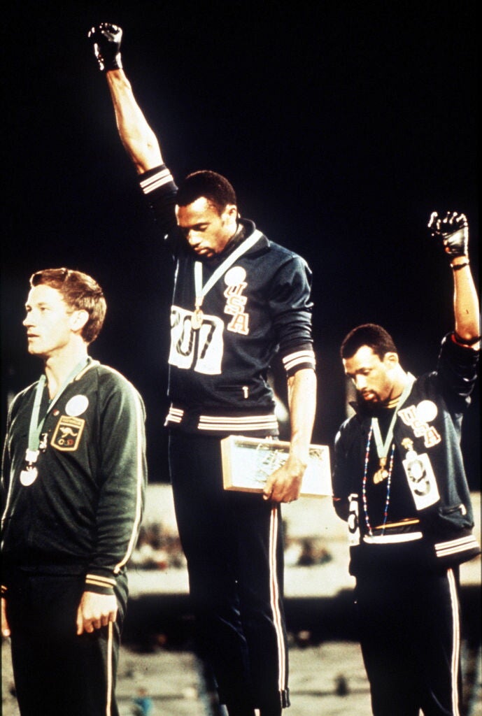 A color photograph of the medals podium at the 1968 Olympic Games. Tommie Smith (the gold medalist, center), a Black man, is wearing a dark blue Team USA jacket and pants, a gold medal around his neck, and his right arm raised, his gloved right hand in a clenched fist. John Carlos (bronze medalist, right), a Black man, is also wearing a Team USA uniform with the bronze medal around his neck. His left arm is raised, his gloved left hand in a clenched fist. Peter Norman (silver medalist, left), a White man, i