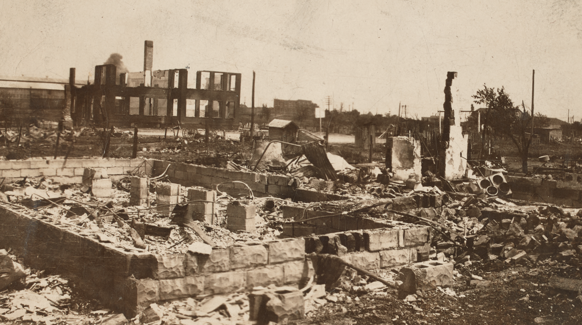 A black-and-white photograph of the destroyed Greenwood District in Tulsa, Oklahoma after the Tulsa Race Massacre. In the foreground is a stone foundation littered with and surrounded by ash. The background features remnants of building façades.