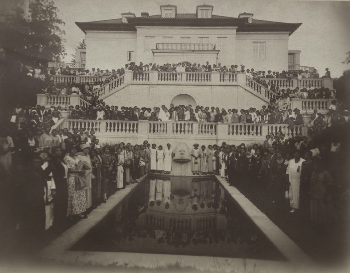 Black and white photograph of a large group of people, mostly women, posed around the exterior of a large Italianate style mansion. The figures are arranged on the building's multi-level balconies, stairs, and around a rectangular pool on the ground level. The photograph is mounted on a piece of gray cardboard that has sustained loss at the top right and bottom right corners. In the bottom right corner is black script that is partially missing.