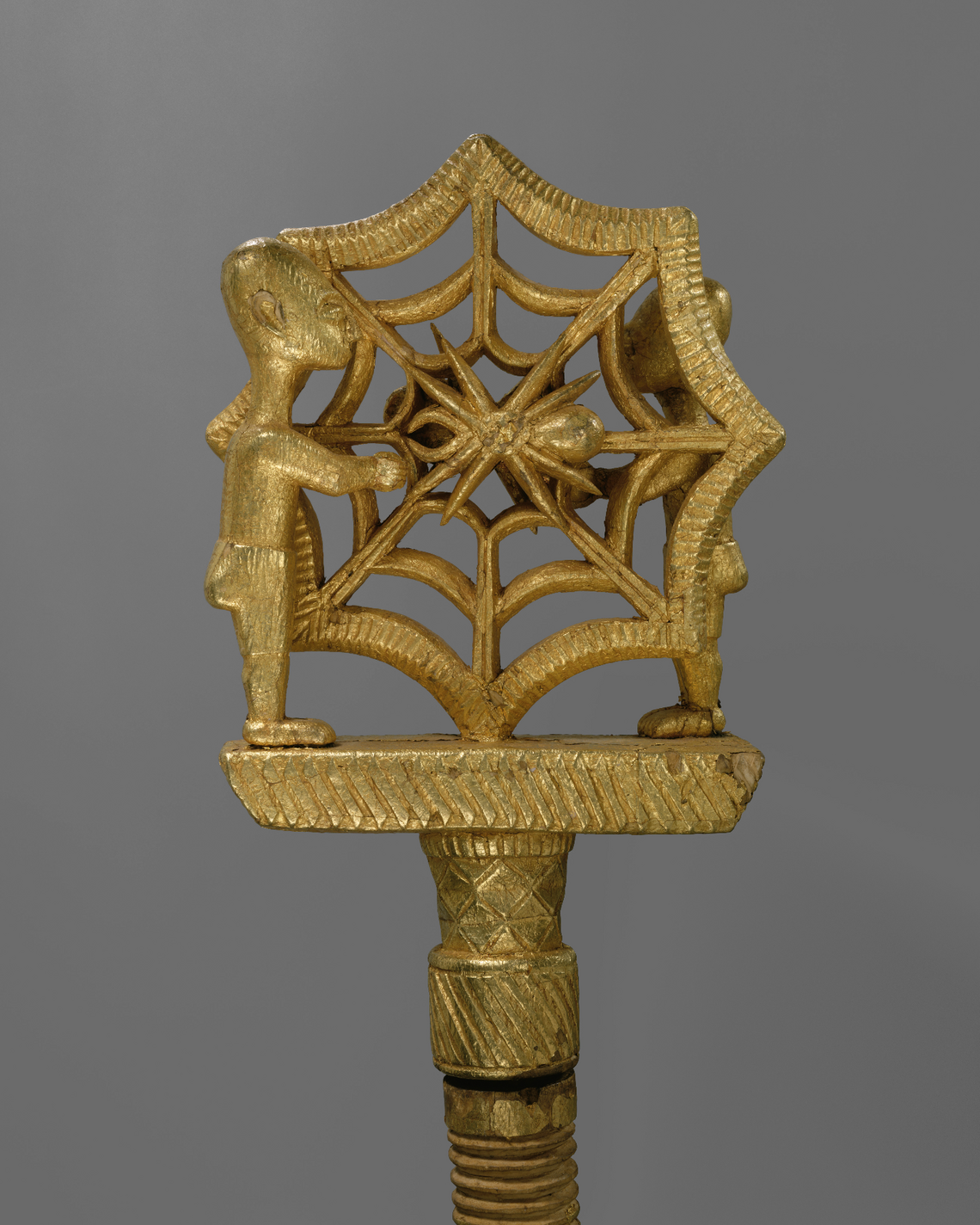 A wooden staff covered with gold leaf depicting two human figures on either side of a spider web and spider