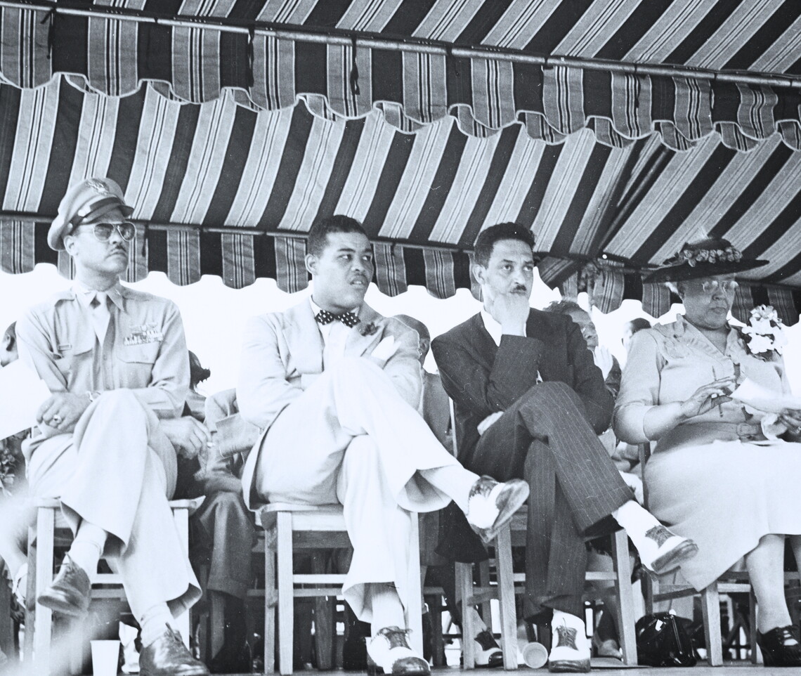 A black-and-white photograph of Benjamin O. Davis Jr. (a Black soldier in uniform), Joe Louis (a Black man wearing a light colored suit and a dark bowtie), Thurgood Marshall (a Black man wearing a dark suit and white shirt), and Daisy Lampkin (a Black woman wearing a light colored dress and dark hat) seated outside under an open-air tent.