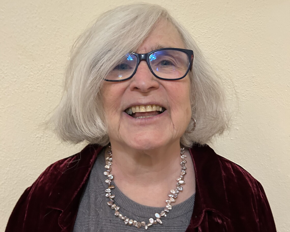 A bespectacled, smiling white woman with white hair wearing a gray shirt, black sweater, and white necklace