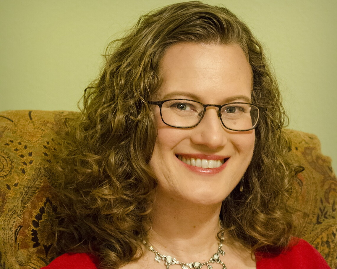 a white woman with long brown hair wearing glasses, a metallic necklace, and a red shirt