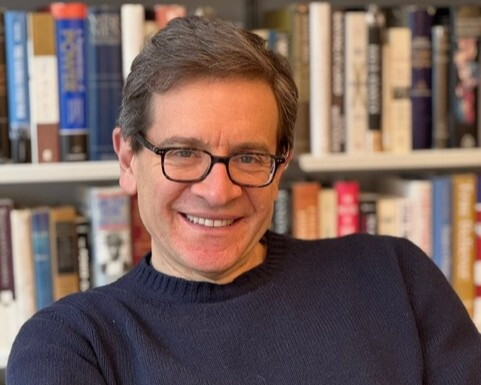 A white man wearing glasses and a dark sweater in front of a bookshelf.