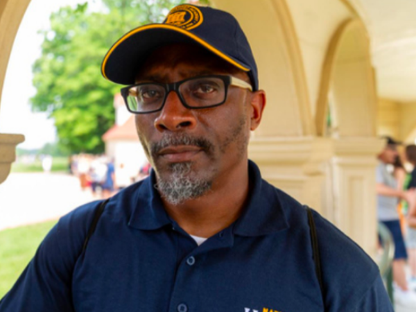 A Black man wearing a dark blue cap with orange piping and a blue polo shirt.